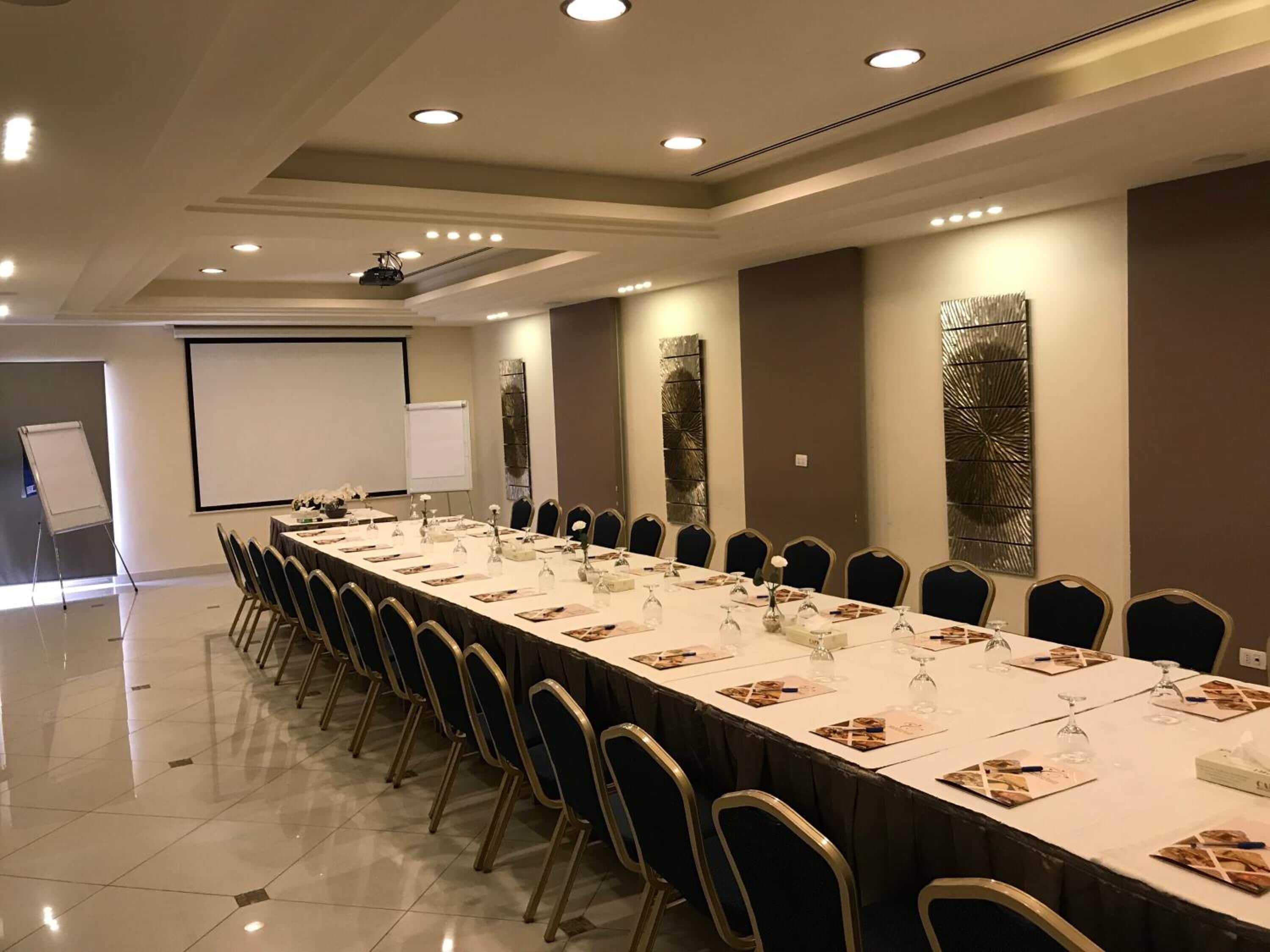 Un salón de conferencias con una larga mesa, sillas, una pantalla de proyector y detalles decorativos en la pared.