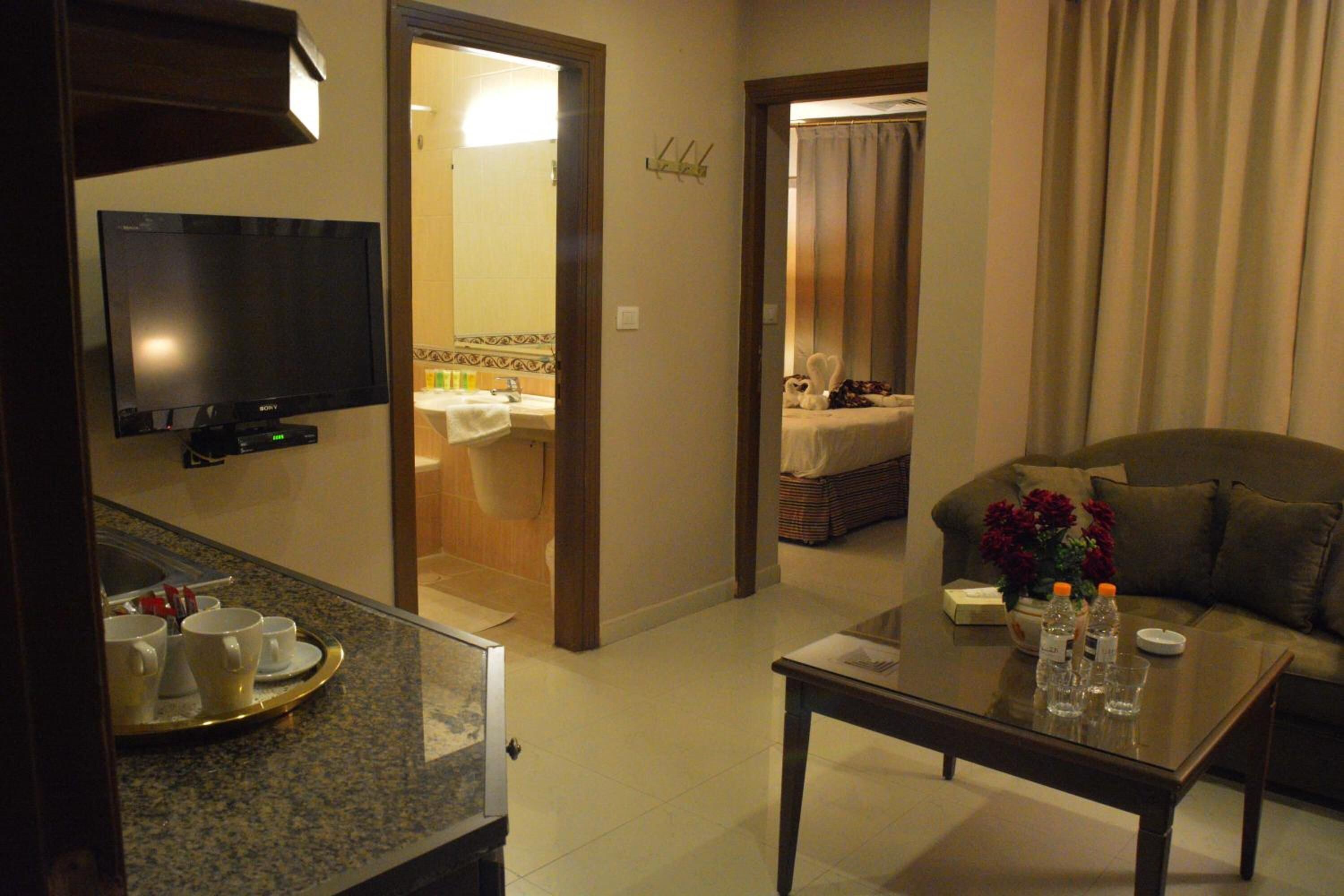 A hotel room with a flat-screen TV, a dining area with a buffet setup, a living room with a sofa and a coffee table, and a bathroom visible through an open door.