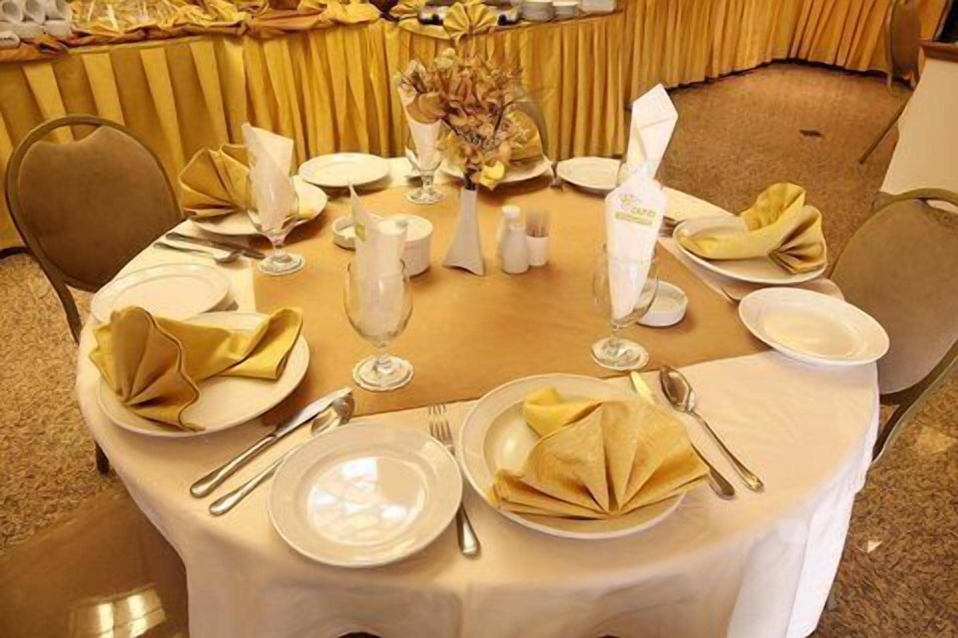 A round dining table set with white plates, gold napkins, and silverware, surrounded by chairs.