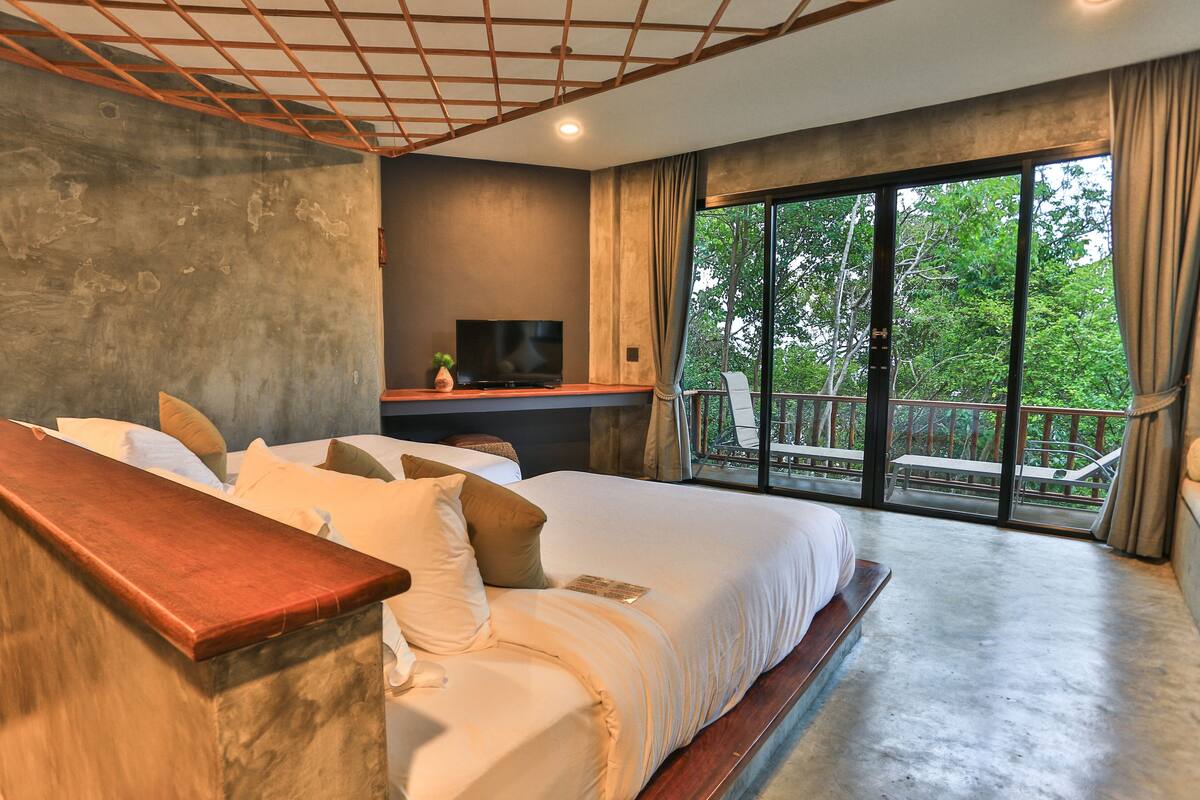 A bedroom with a large bed, a desk with a TV, and a balcony with a view of trees.