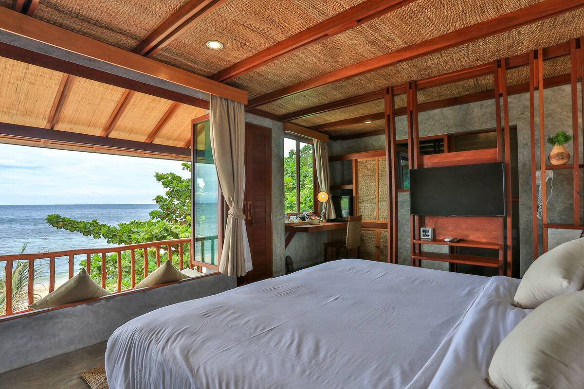 A bedroom with a large bed, a desk with a computer, a TV, and a view of the ocean.