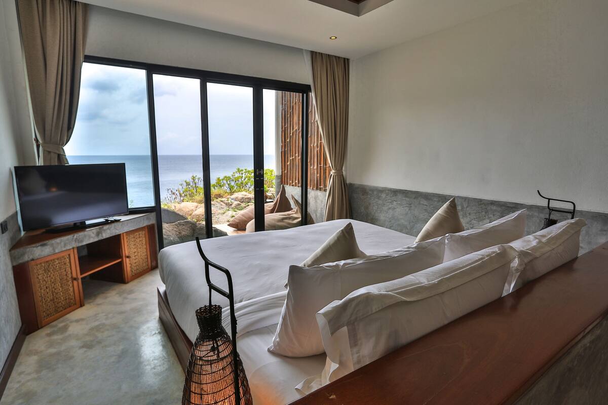 A bedroom with a large bed, a TV on a stand, and a view of the sea.