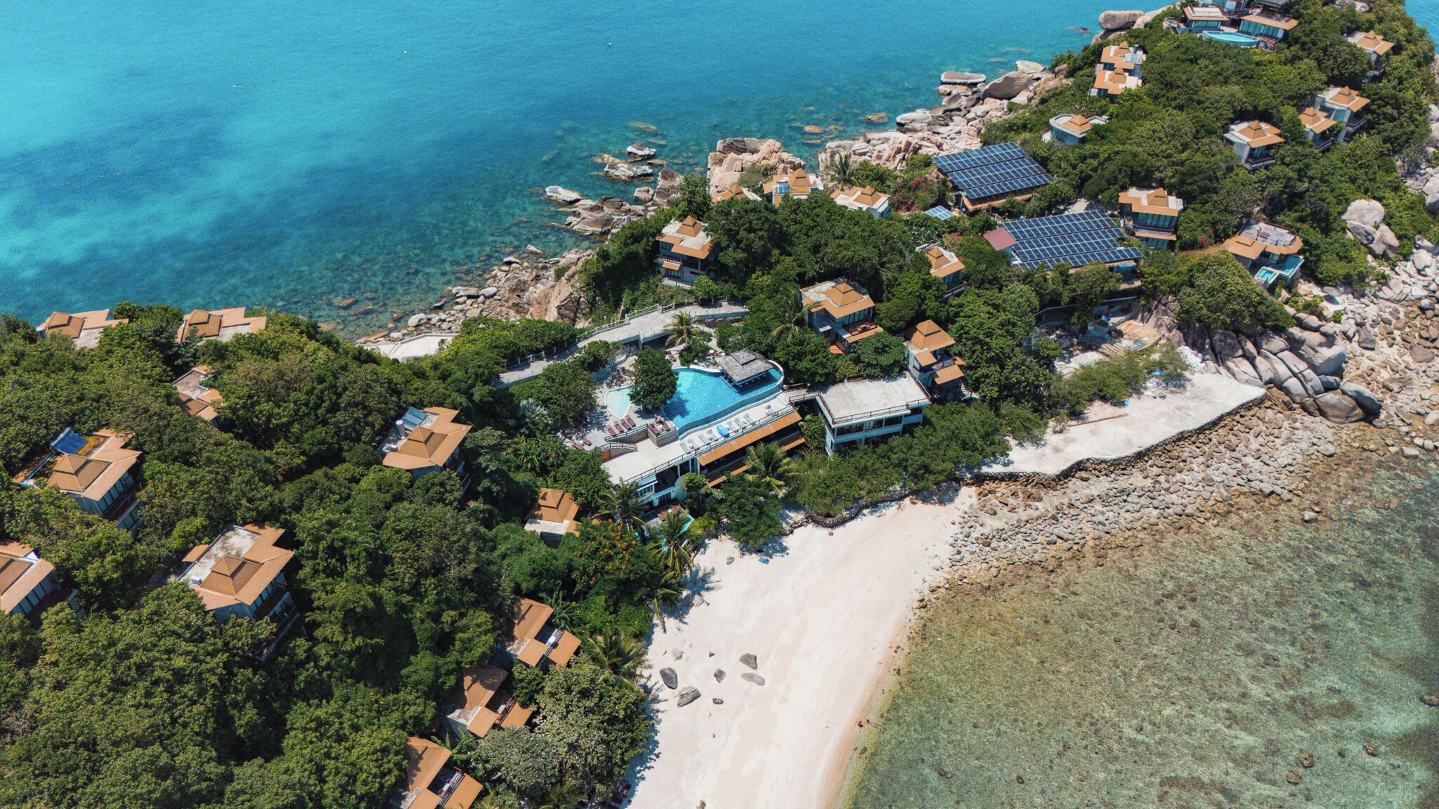 An aerial view of a coastal resort with multiple villas, a central swimming pool, and a beach.