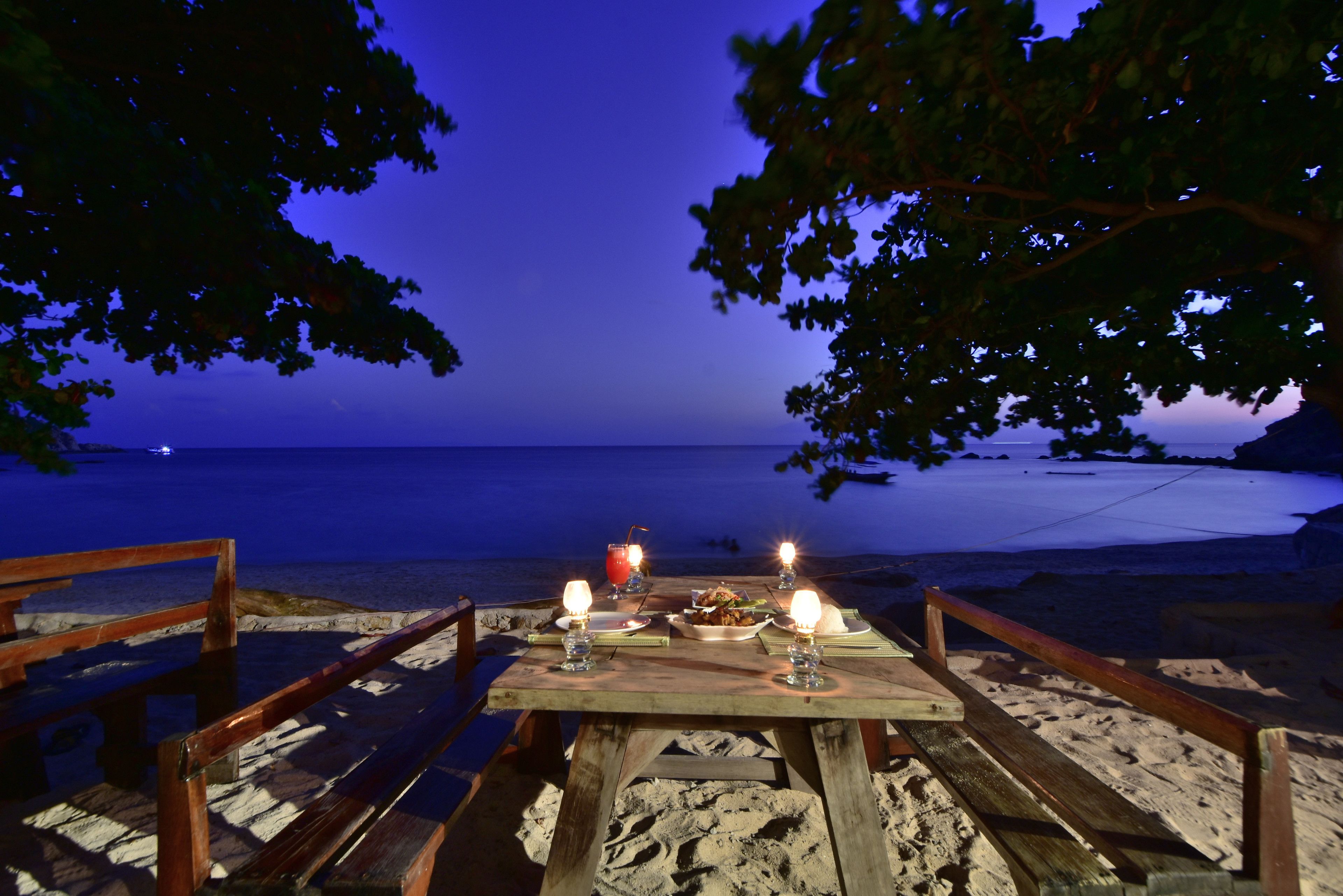 A wooden table with candles and a drink, set on a sandy beach with a view of the ocean.