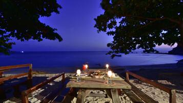 A wooden table with candles and a drink, set on a sandy beach with a view of the ocean.