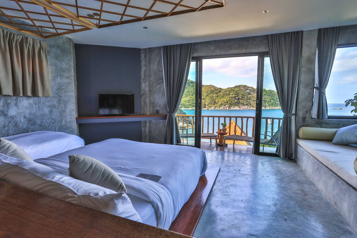 A bedroom with a large bed, a TV on a stand, and a balcony with a view of the sea.