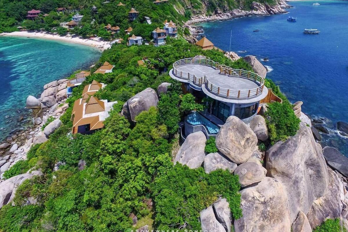 A luxurious cliffside resort with a circular building featuring a rooftop terrace, surrounded by lush greenery and overlooking a clear blue sea.