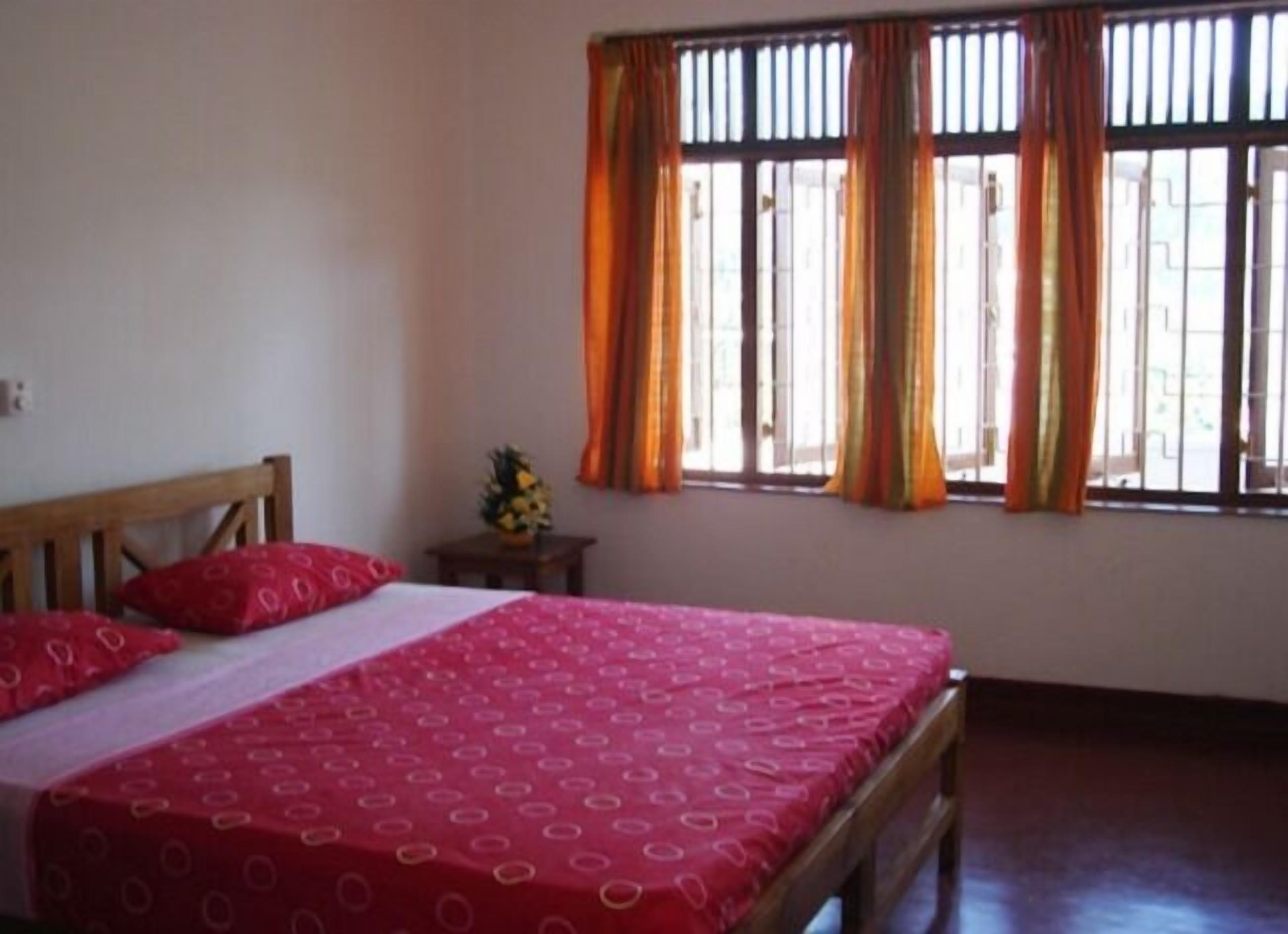 A bedroom with a wooden bed, red bedding, a small table with a pineapple, and a window with orange curtains.