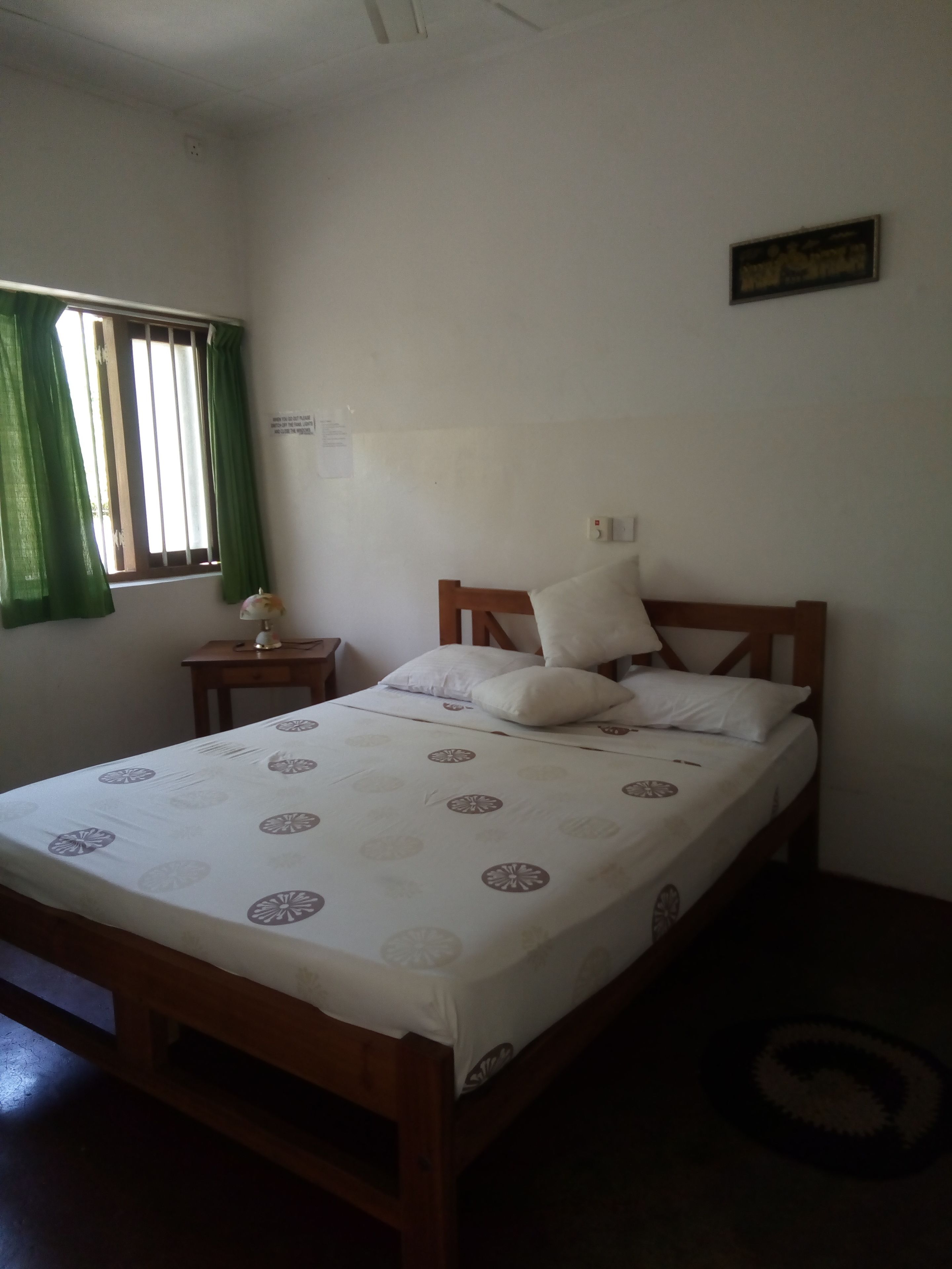 Habitación individual con cama de estructura de madera, colcha blanca con diseño circular, mesita de noche con lámpara, ventana con cortinas verdes y un interruptor de luz en la pared.