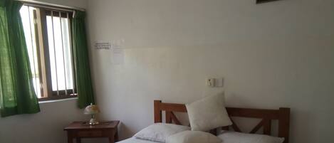 Habitación individual con cama de estructura de madera, colcha blanca con diseño circular, mesita de noche con lámpara, ventana con cortinas verdes y un interruptor de luz en la pared.