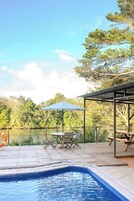 Un área de piscina con cubierta de madera, sillas de descanso y una zona de estar techada con una sombrilla.