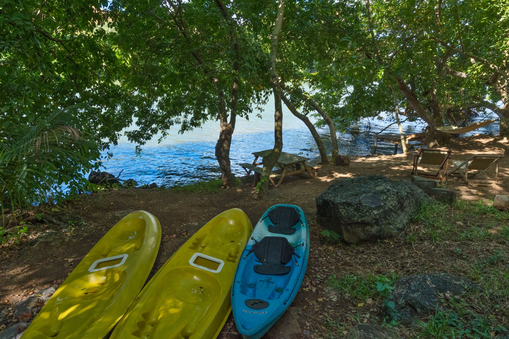 Two kayaks and a hammock near a body of water, with trees and a bench in the background.