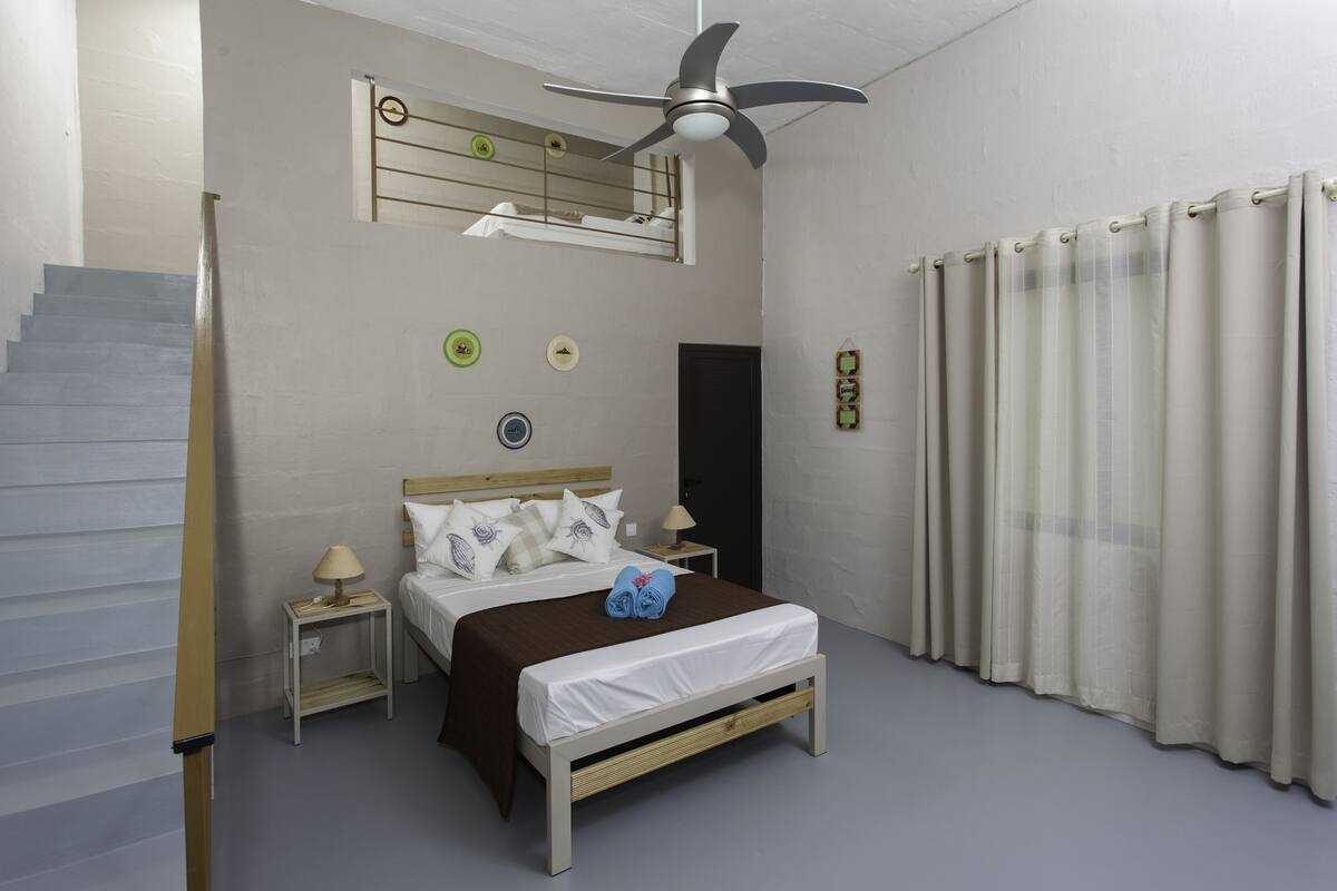 Een slaapkamer met een bed, nachtkastje, wandklok, plafondventilator en een raam met gordijnen.