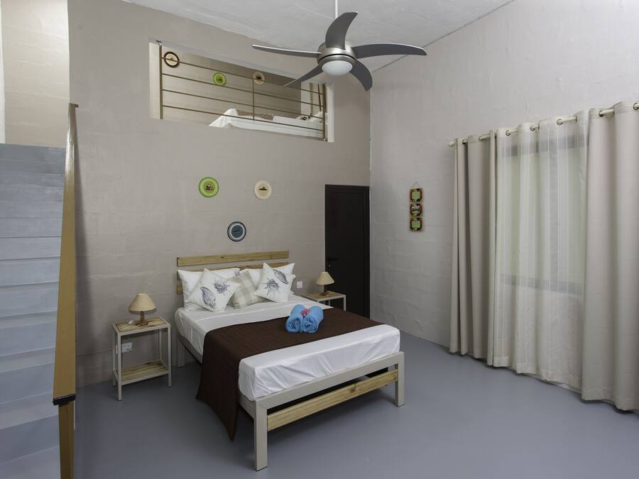 Un dormitorio con cama, mesita de noche, reloj de pared, ventilador de techo y ventana con cortinas.