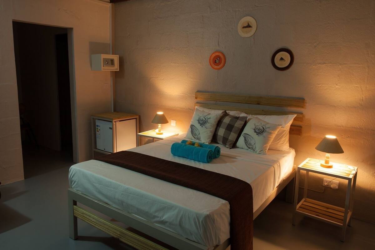 Een net opgemaakt bed met een bruin hoofdeinde, twee nachtkastjes met lampen en een wandklok.