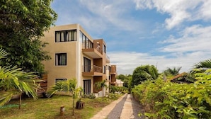 A modern multi-story building with balconies, surrounded by lush greenery and a clear sky.