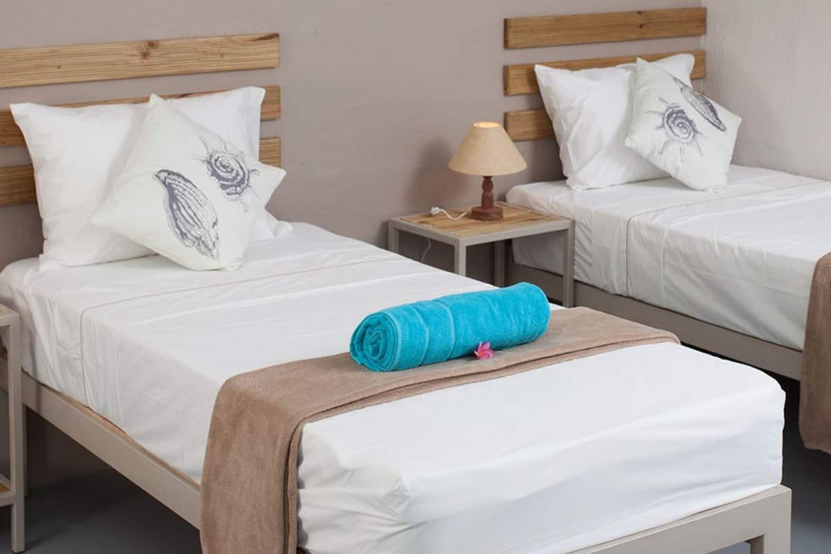 Een hotelkamer met twee eenpersoonsbedden, elk met een houten hoofdbord, witte beddengoed en een blauwe handdoek. Er zijn nachtkastjes met lampen en een stopcontact.