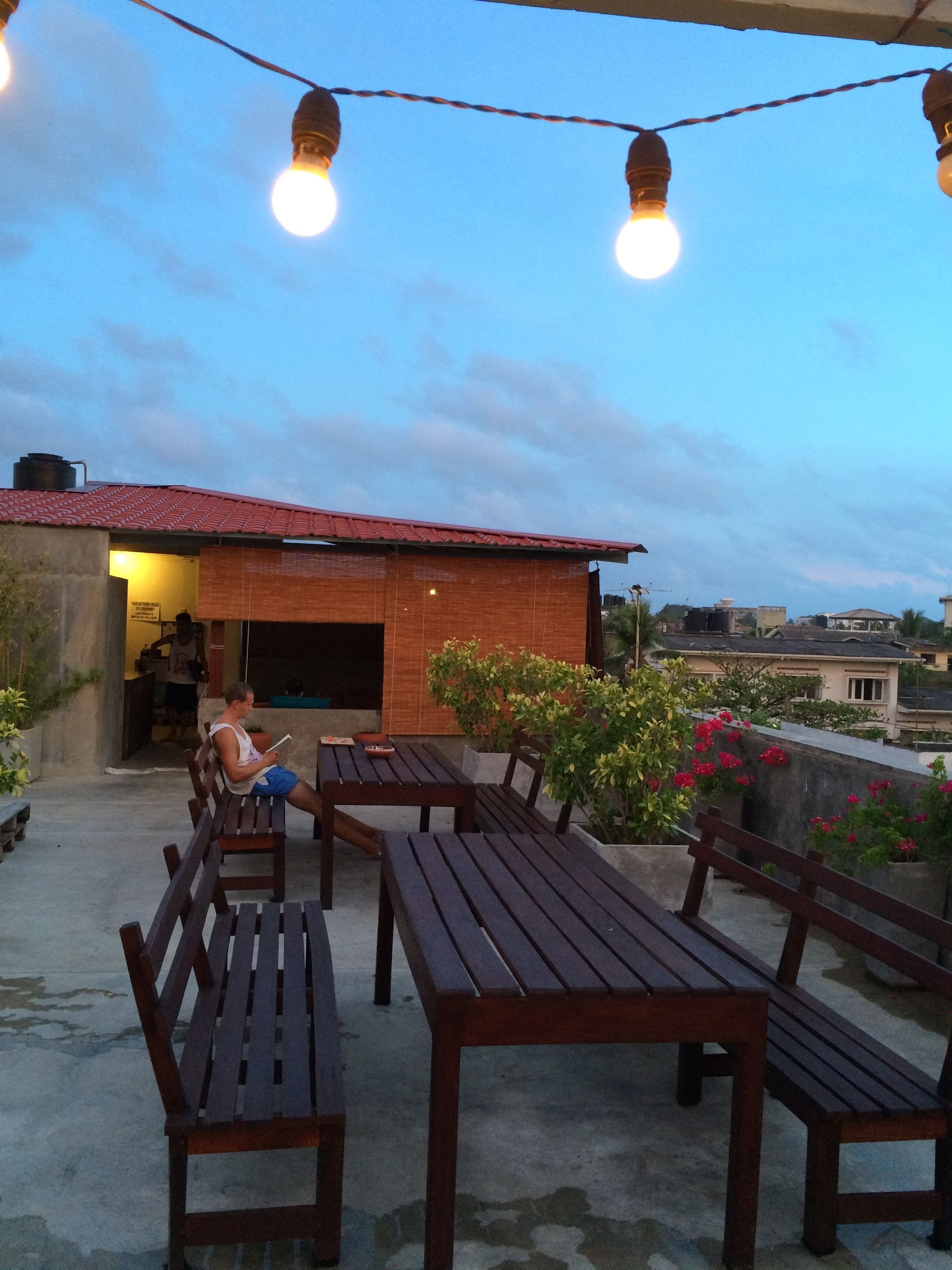 An outdoor seating area with wooden benches and tables, string lights overhead, and a view of the surrounding buildings.