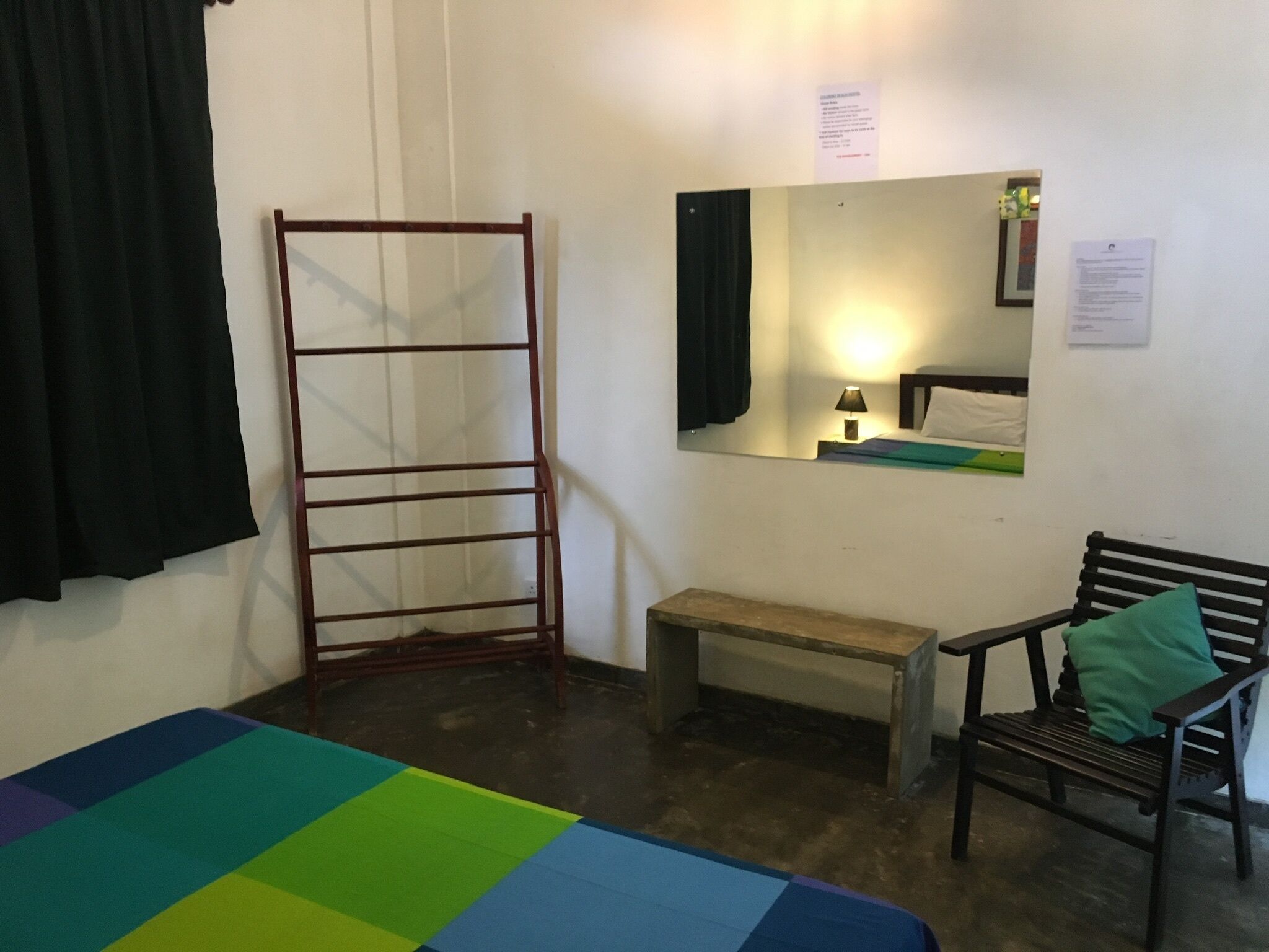A small, simple room with a bed, a bench, a chair, a metal rack, and a mirror on the wall.