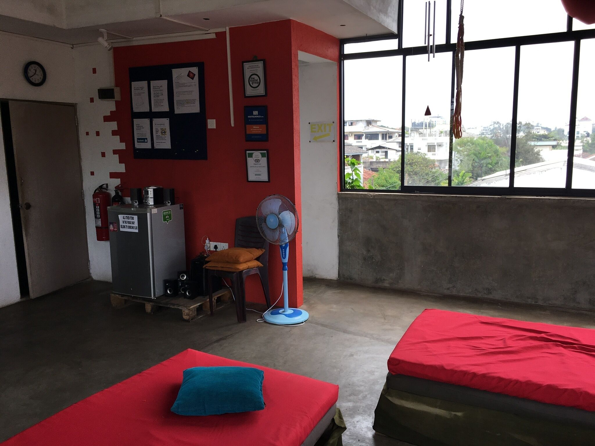 Una habitación con dos camas, una pared roja, un ventanal con vistas, un ventilador y una pequeña mesa con una silla.