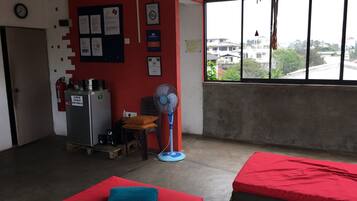 Una habitación con dos camas, una pared roja, un ventanal con vistas, un ventilador y una pequeña mesa con una silla.