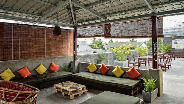 A covered outdoor seating area with a sofa, cushions, a wooden table, and a wicker chair. - Colombo Beach Hostel (Mount Lavinia)