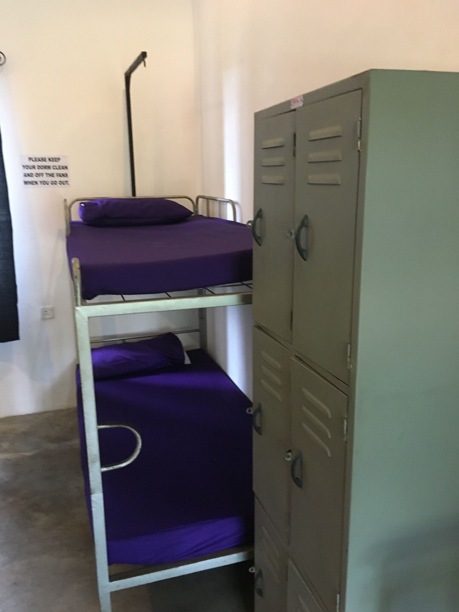A bunk bed with purple bedding and a metal frame, a green metal locker, and a sign on the wall.