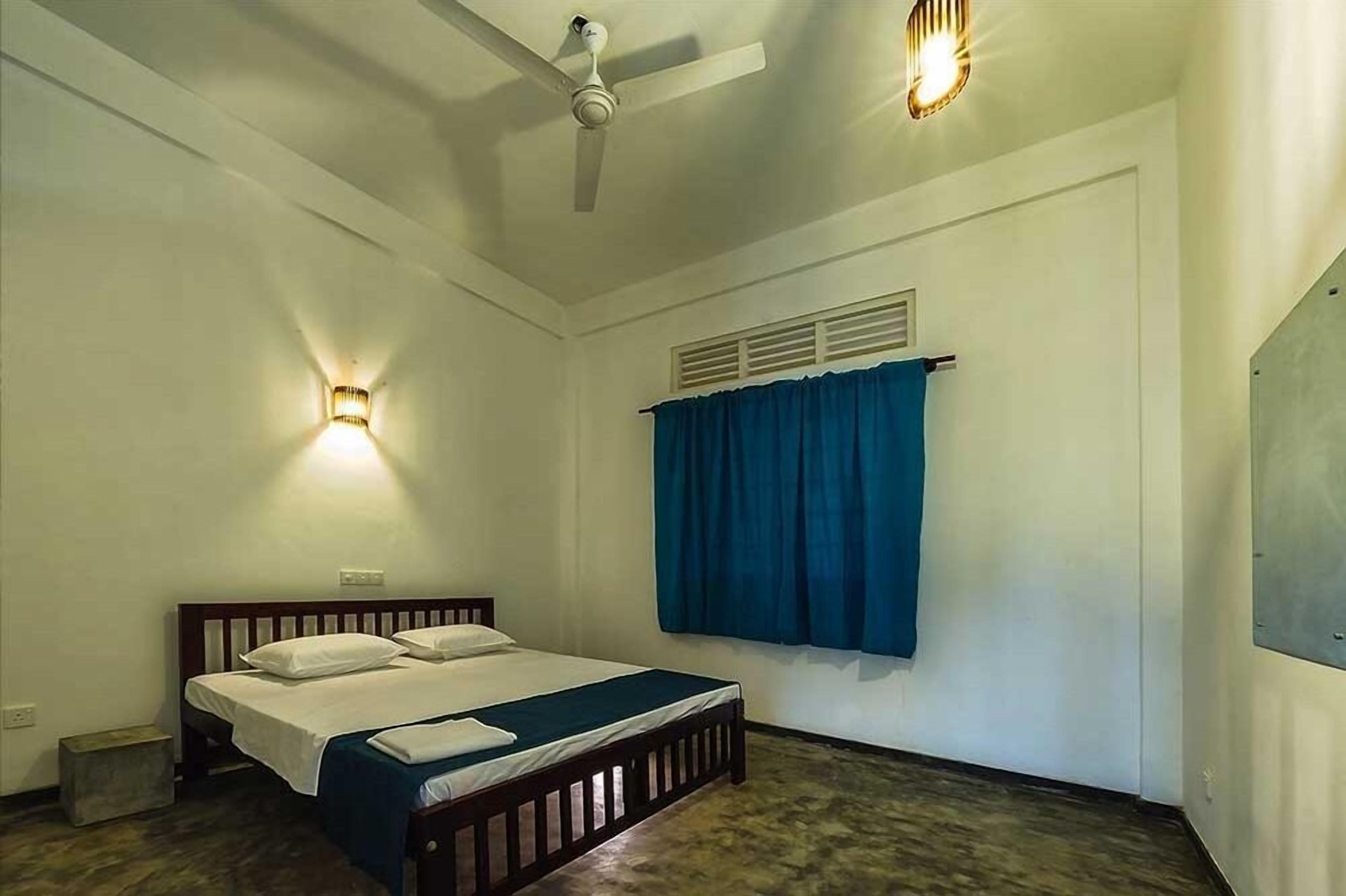 Un lit simple avec un cadre en bois, un ventilateur de plafond et deux lampes fixées au mur.