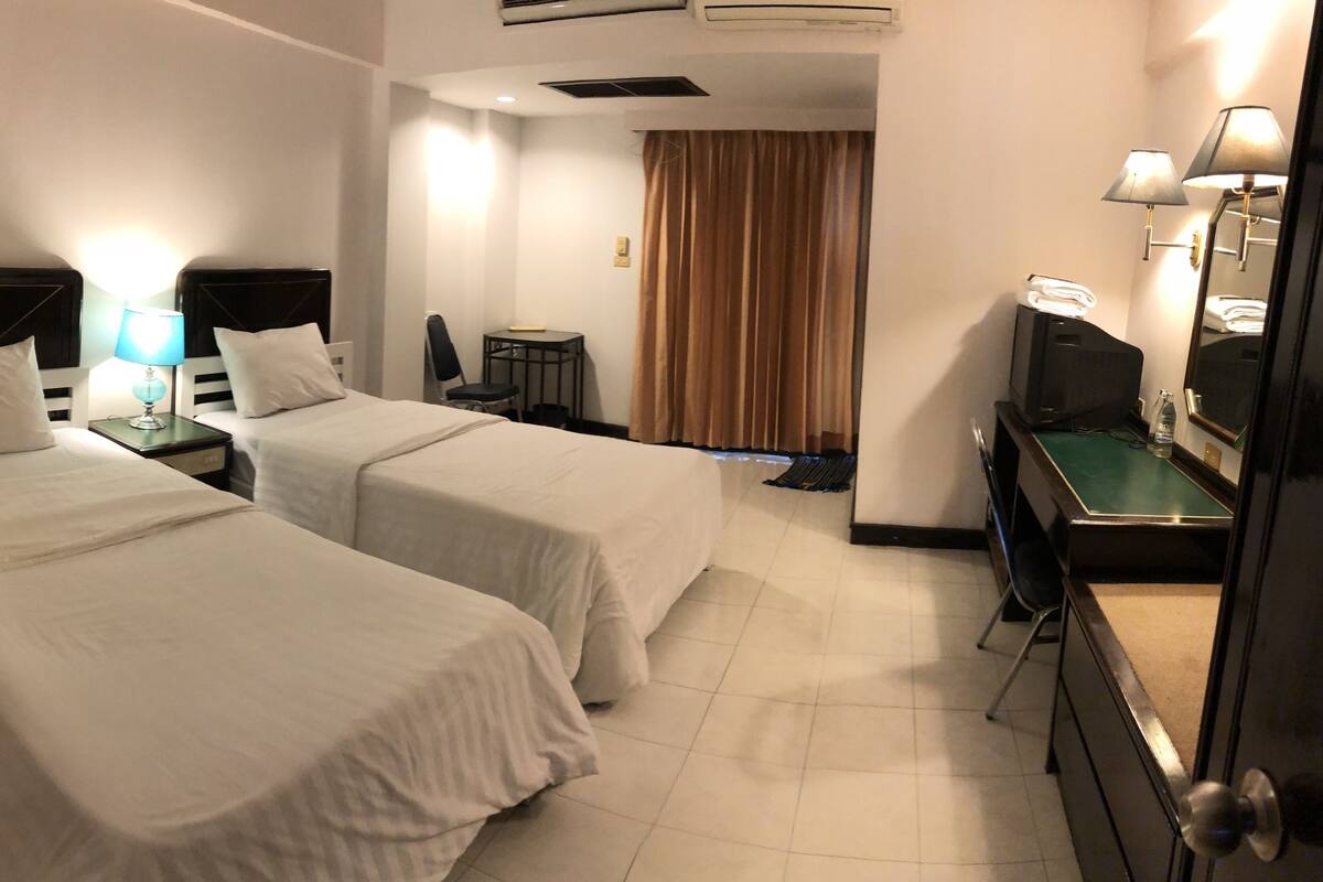 A hotel room with two single beds, a desk with a computer, a chair, and a bathroom visible in the background.