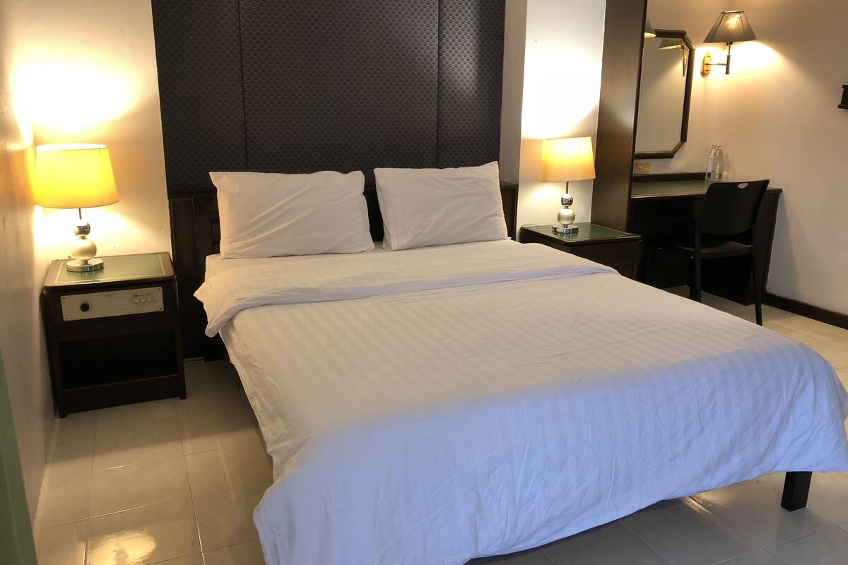 A neatly made bed with white linens, two bedside tables with lamps, a chair, and a large mirror on the wall.