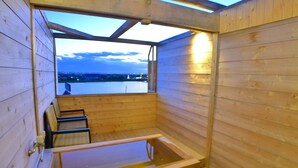 A wooden sauna with a glass door, a bench, and a view of a body of water and mountains. - New Makomo Hotel (Itako)
