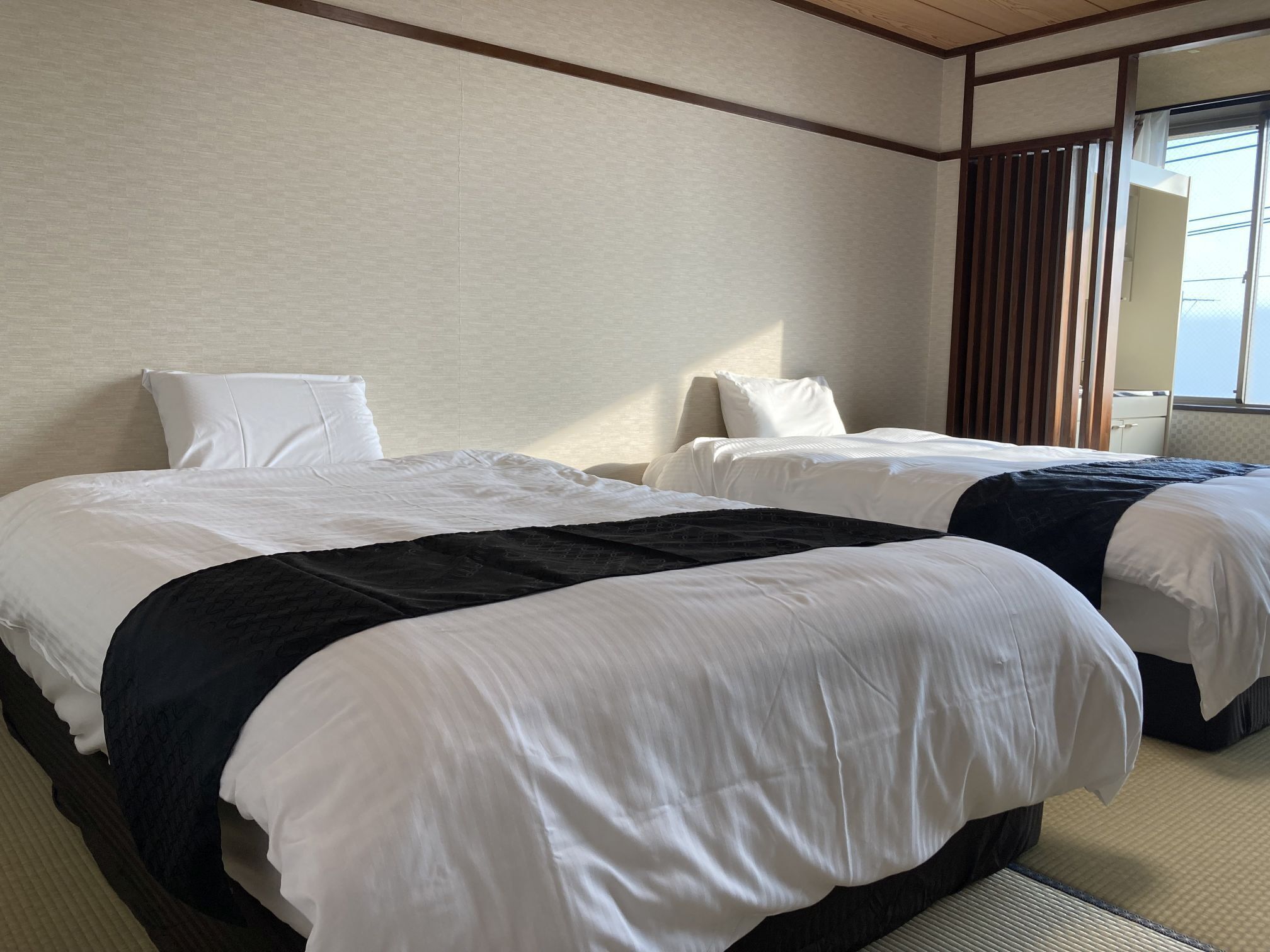 A hotel room with two beds, each with white bedding and a dark blanket. The room has a wooden ceiling and a window with blinds.