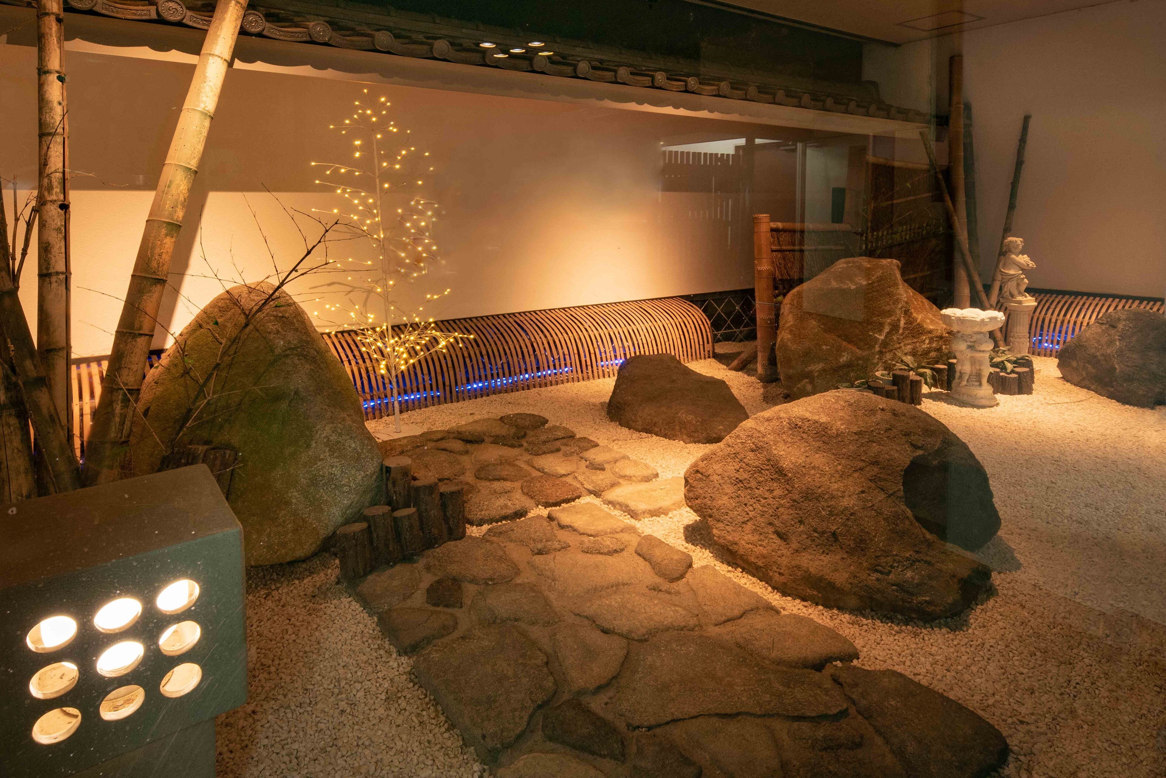 Un jardin japonais traditionnel avec des allées pavées de pierres, de gros rochers, un bambou et une statue.