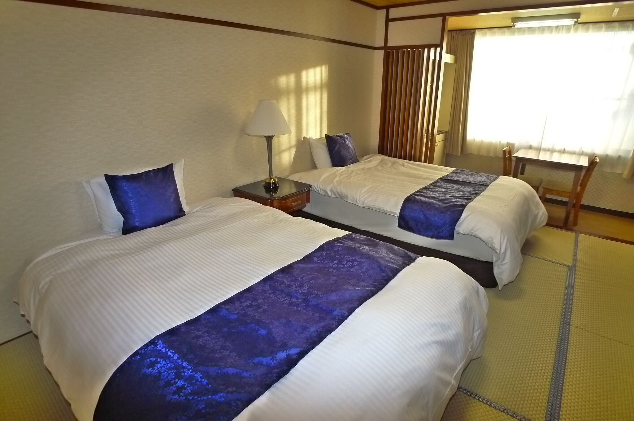 A hotel room with two beds, a nightstand, a lamp, and a window with curtains.