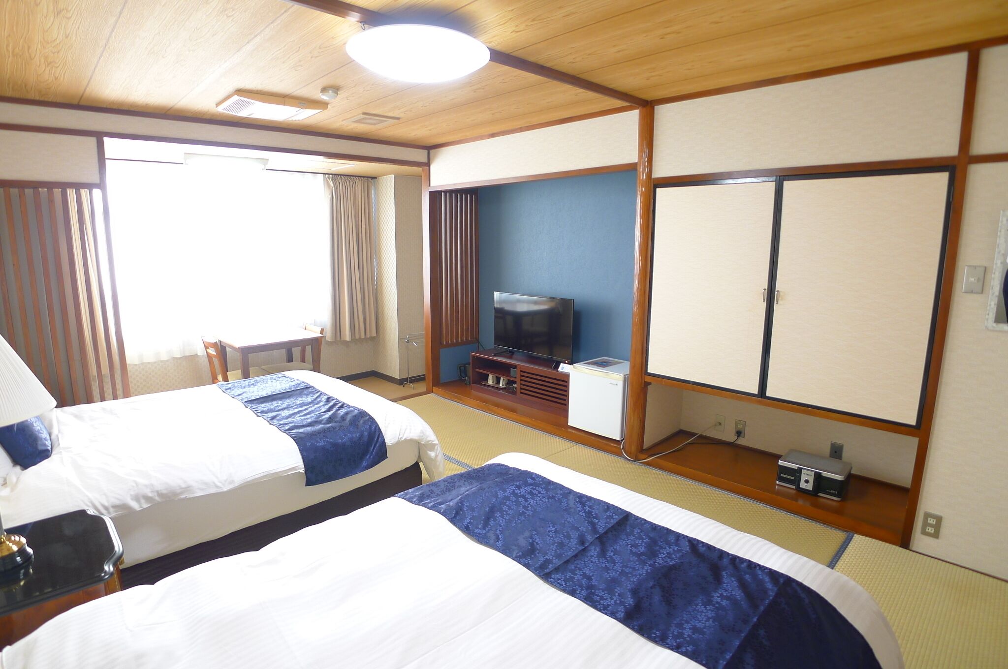 A hotel room with two beds, a television, a small desk, and a window with curtains.