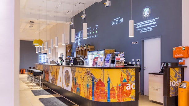 A hotel reception area with a counter, bar stools, a digital clock, and a large wall clock. - a&o Hostel Salzburg Hauptbahnhof (Salzburg)