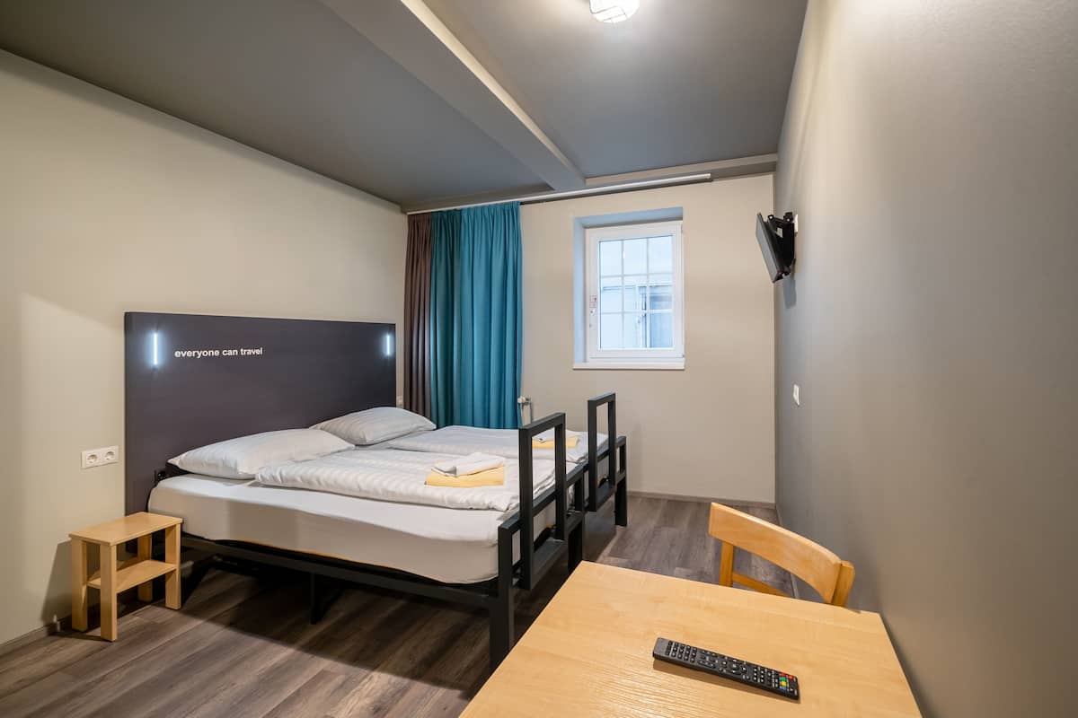 Et lille hotelværelse med en enkelt seng, et lille bord, en stol, et vindue med gardiner og et TV på væggen.