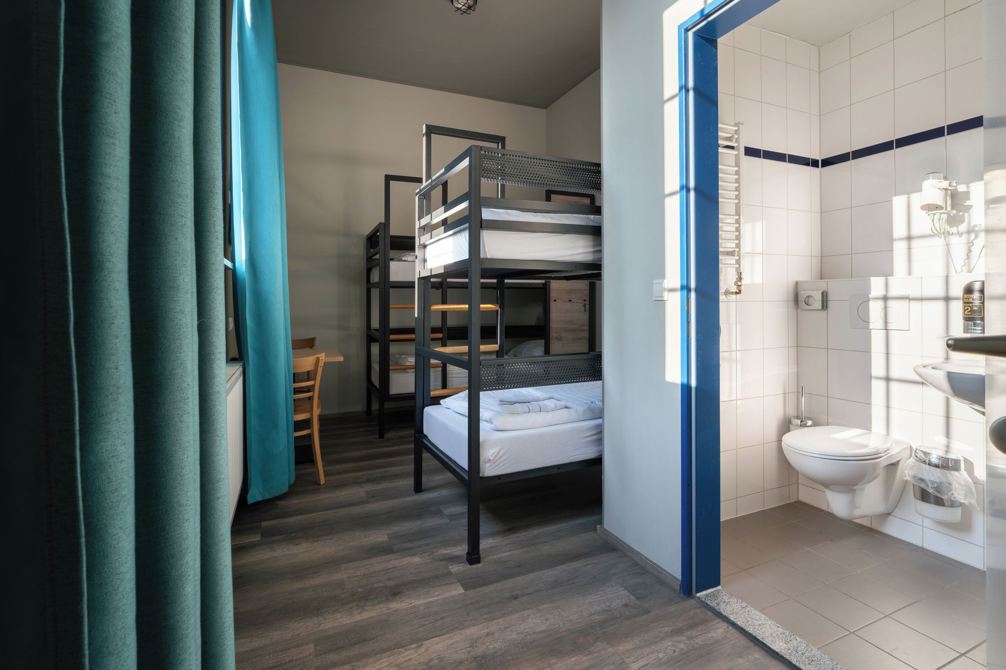 Habitación pequeña con literas, una mesita, una silla y un baño con inodoro y lavabo.