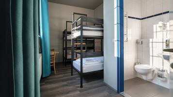 Habitación pequeña con literas, una mesita, una silla y un baño con inodoro y lavabo.