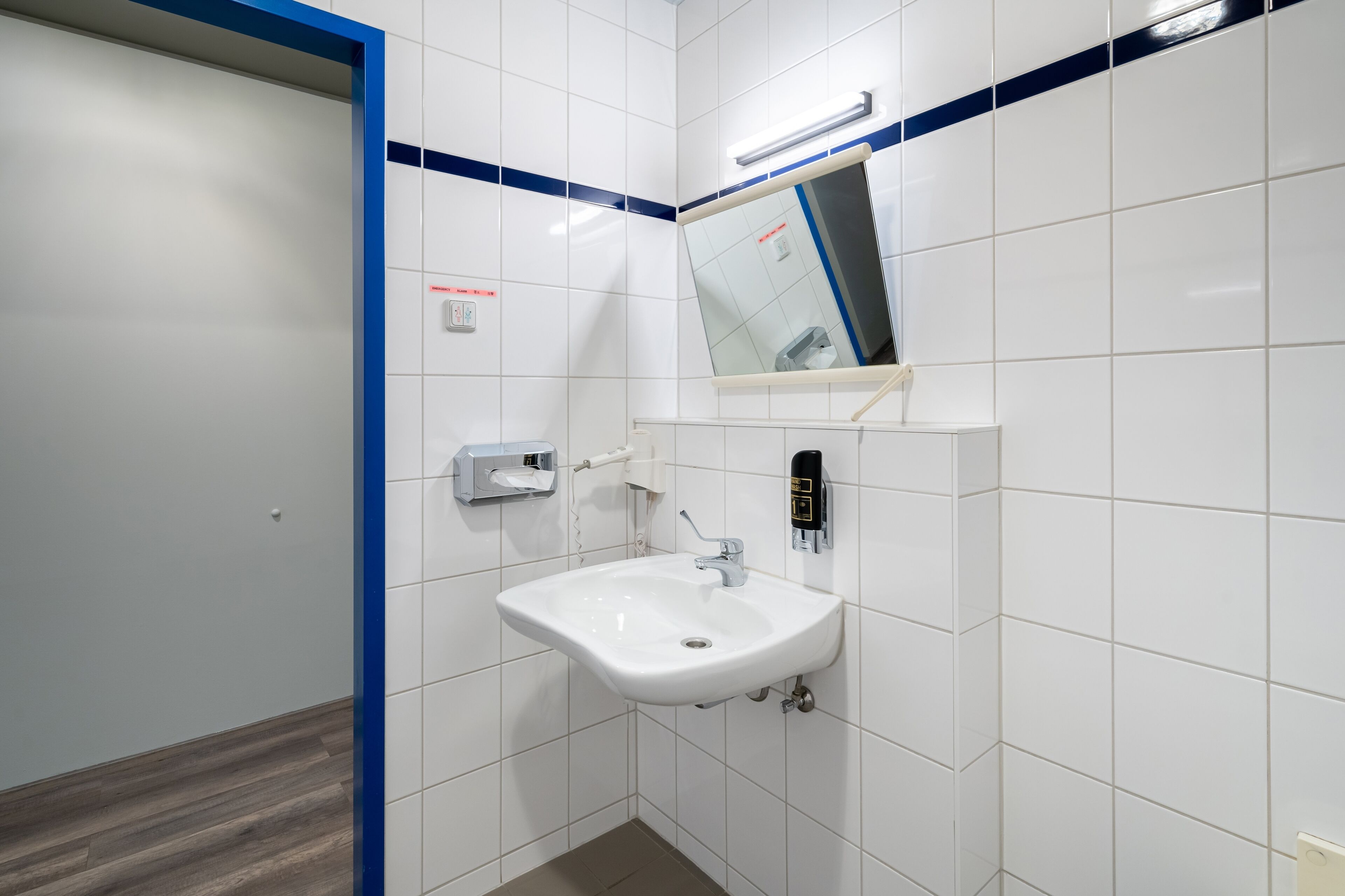 Ein Badezimmer mit weißen Fliesen, einem Waschbecken, einem Spiegel und einer blauen Akzentwand.