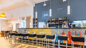 A bar area with stools, a counter with a QR code, shelves with bottles, and a colorful mural. - a&o Hostel Salzburg Hauptbahnhof (Salzburg)