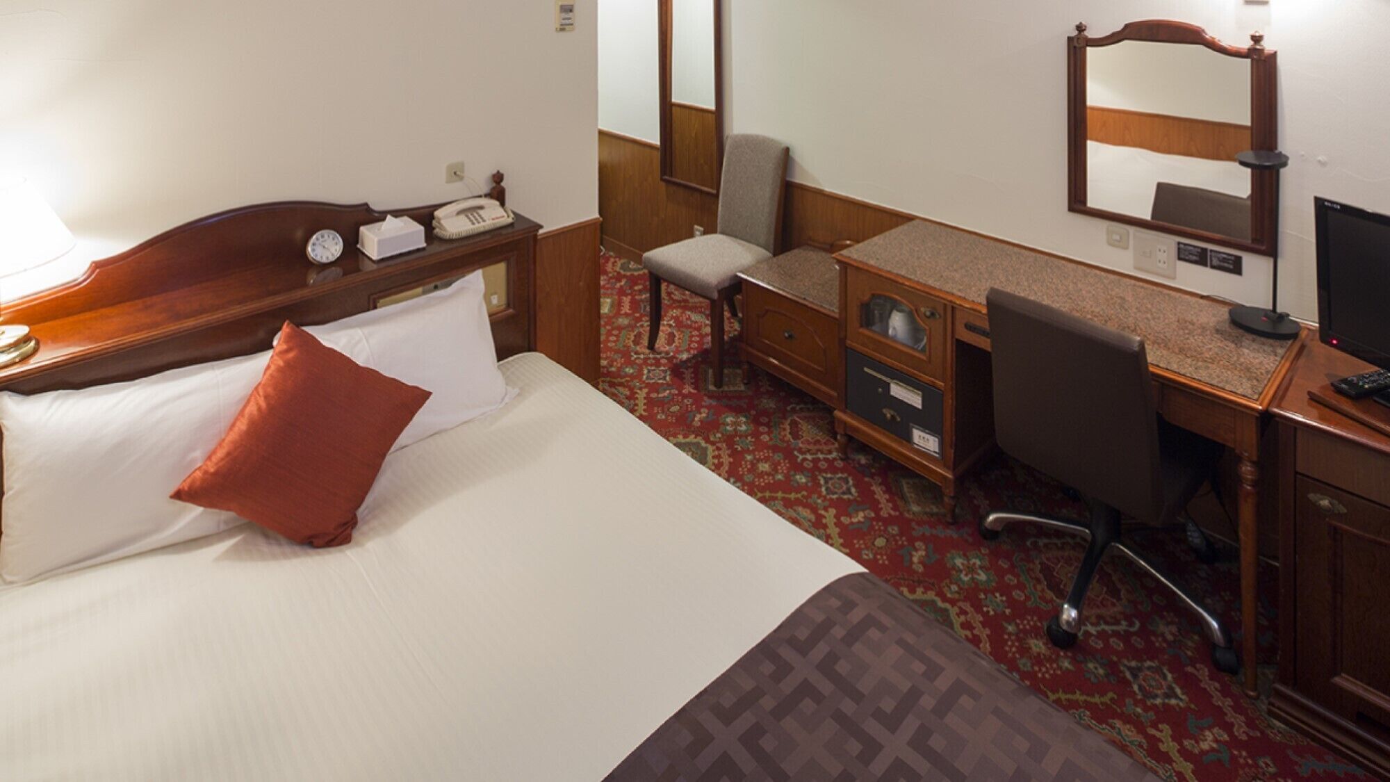 A hotel room with a bed, a desk, a chair, a desk lamp, a telephone, and a television.