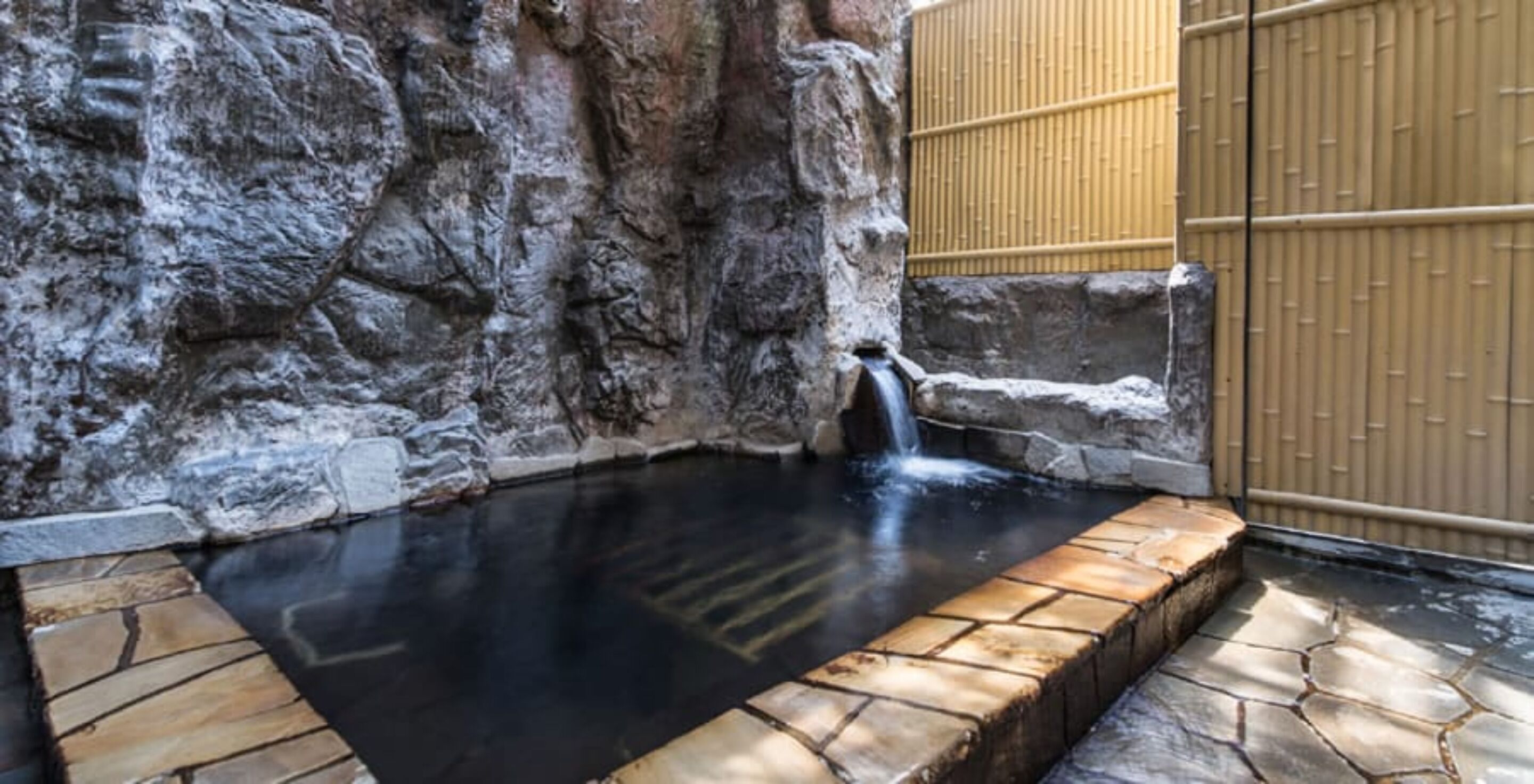 A hot spring area with a stone pool, a waterfall, and a bamboo fence.