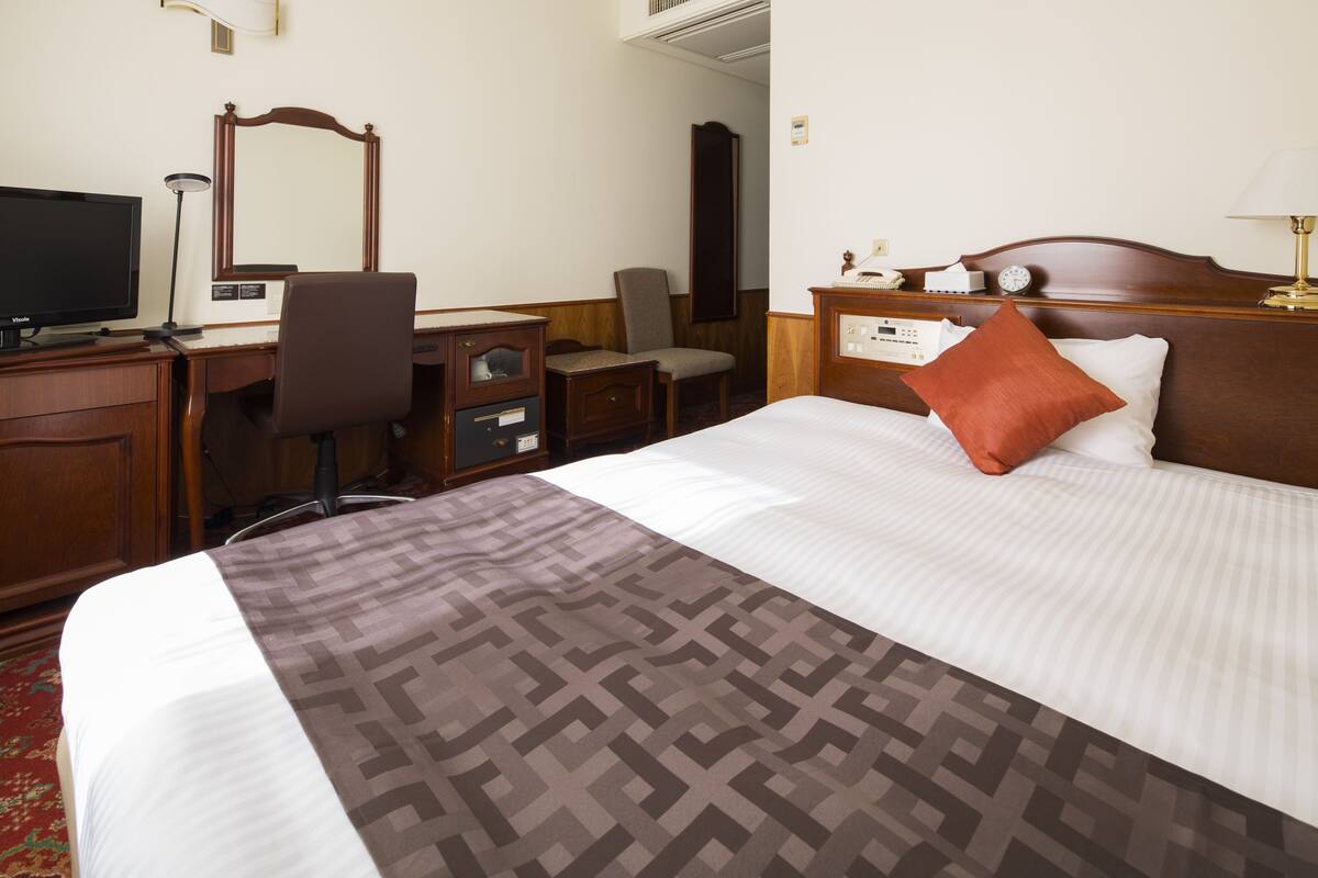 A hotel room with a bed, a desk with a computer, a chair, a mirror, and a bedside table with a lamp.