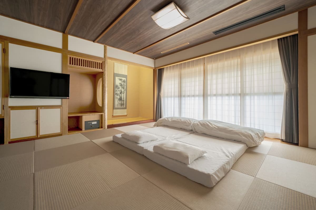 A room with tatami flooring, a bed, a television, and a large window with curtains.