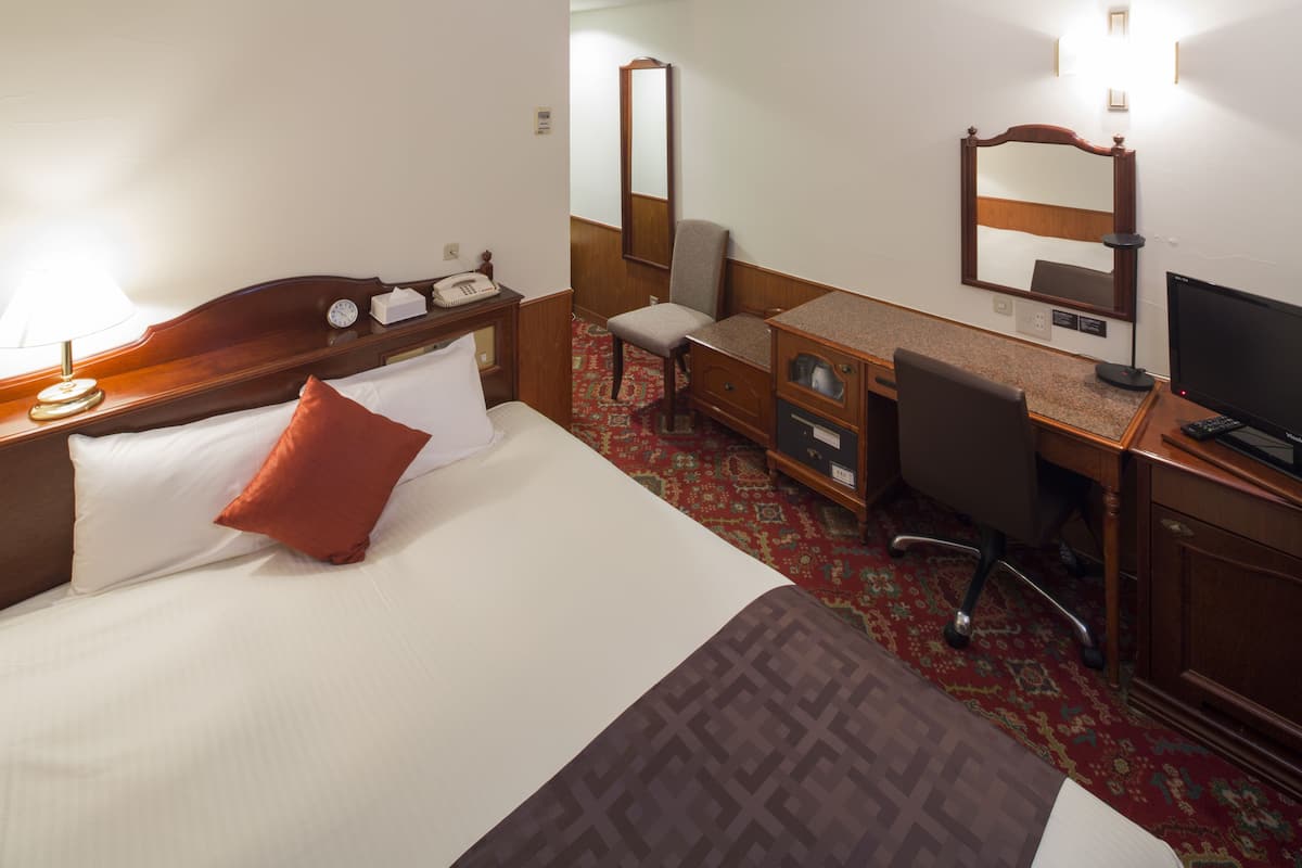 A hotel room with a bed, a desk with a computer, a chair, a dresser, and a mirror.