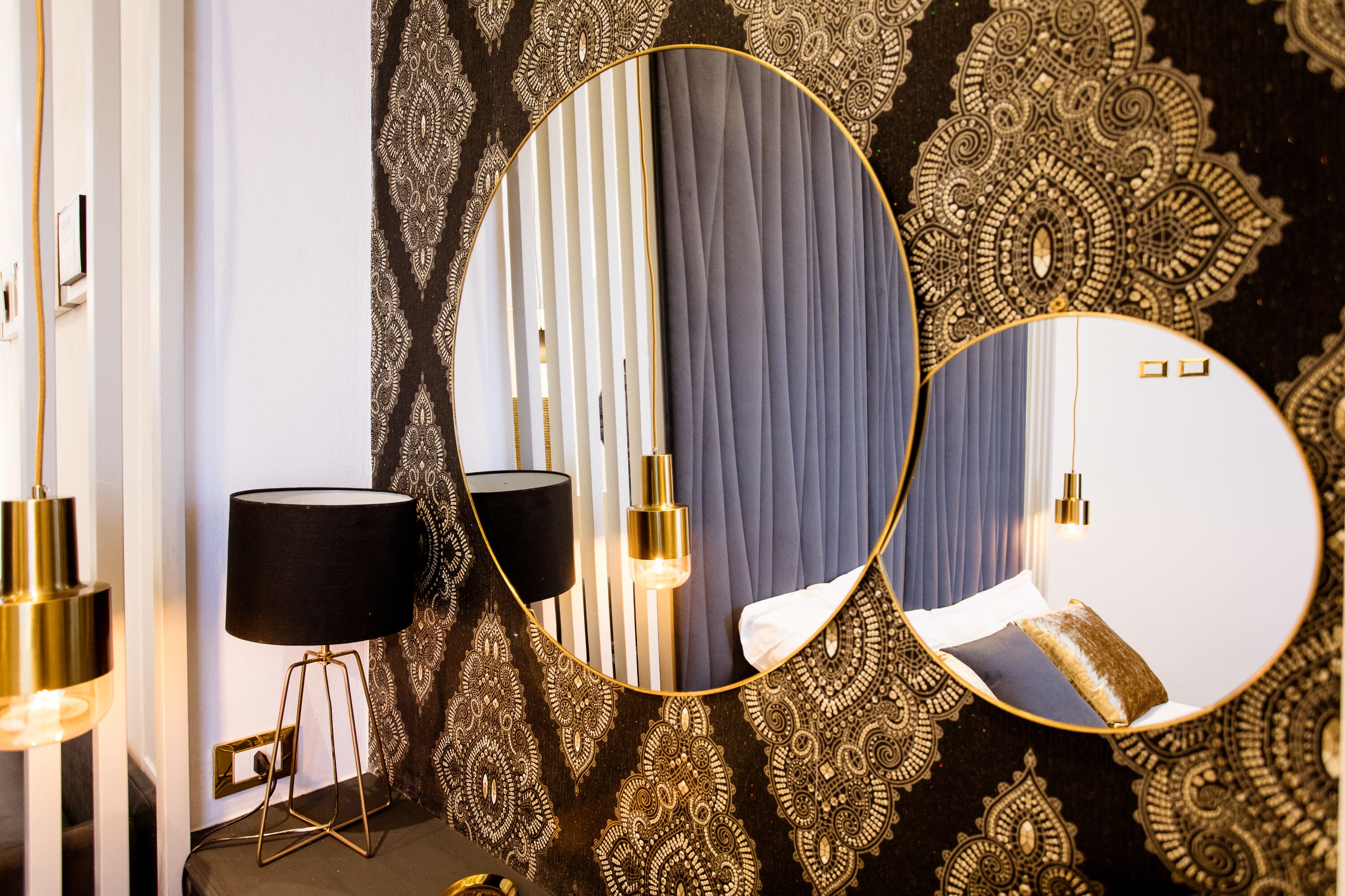 A room with a patterned wallpaper, a round mirror, and a lamp on a bedside table.