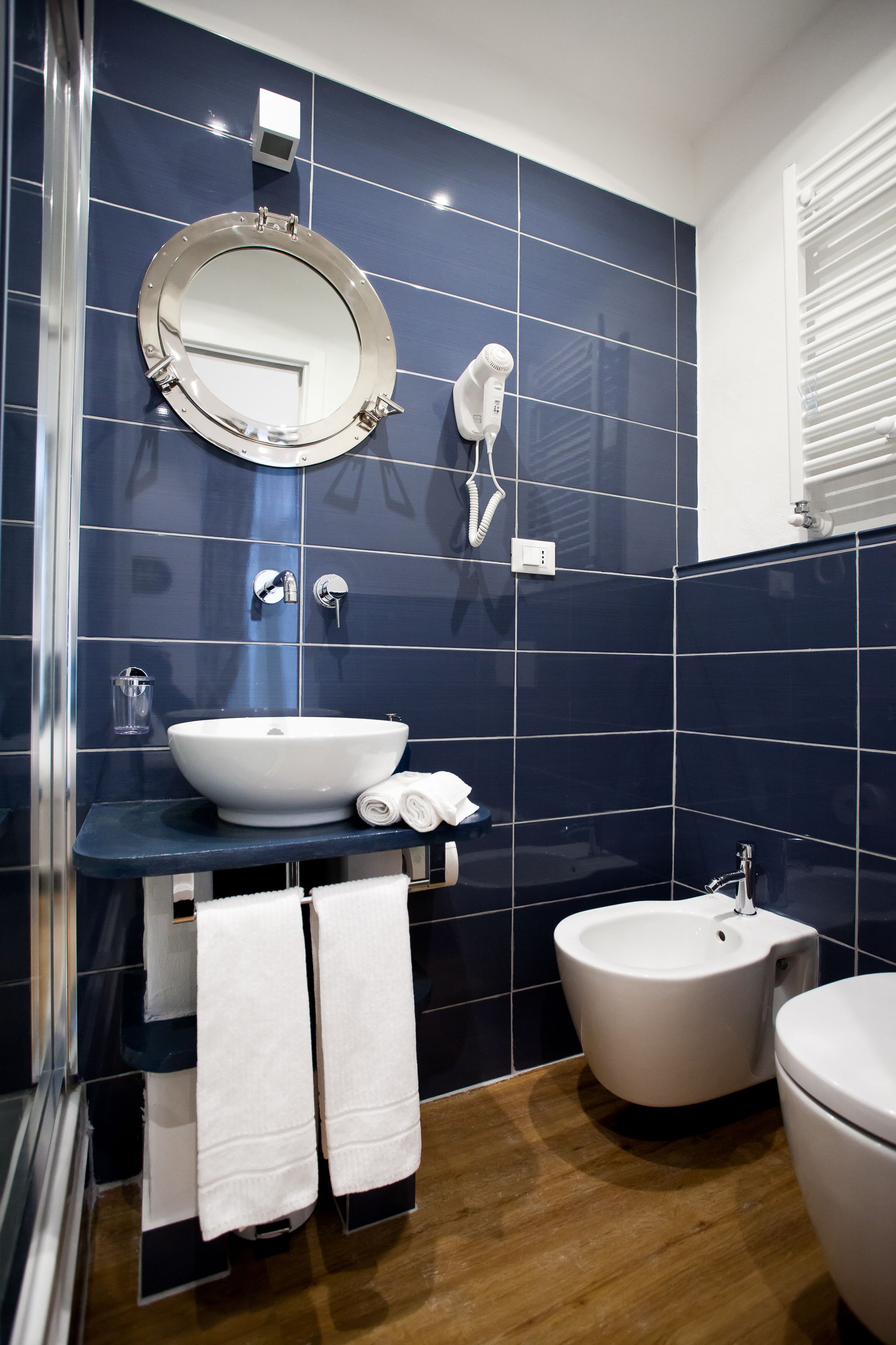 Une salle de bain avec un miroir rond, un lavabo blanc, un bidet et deux serviettes posées sur une étagère.