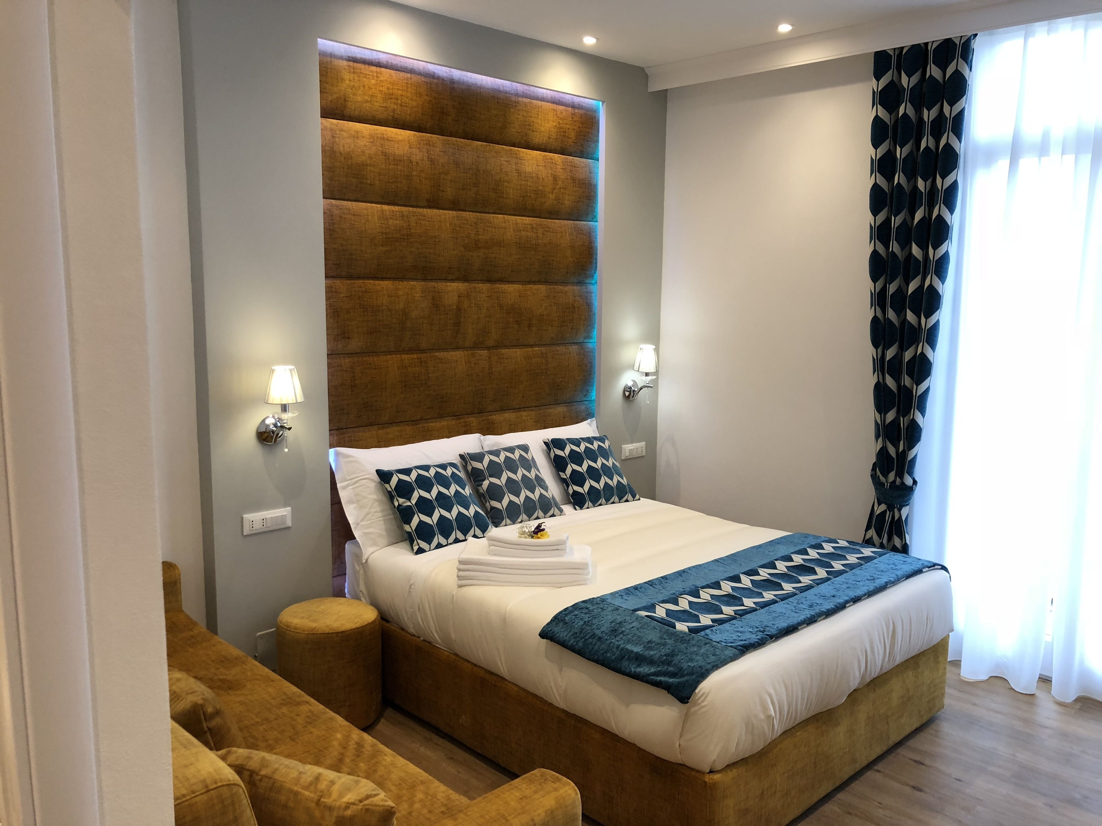 Habitación de hotel moderna con una cama grande, cabecera con iluminación integrada, dos lámparas de noche, una manta decorativa y vistas al exterior a través de cortinas traslúcidas.