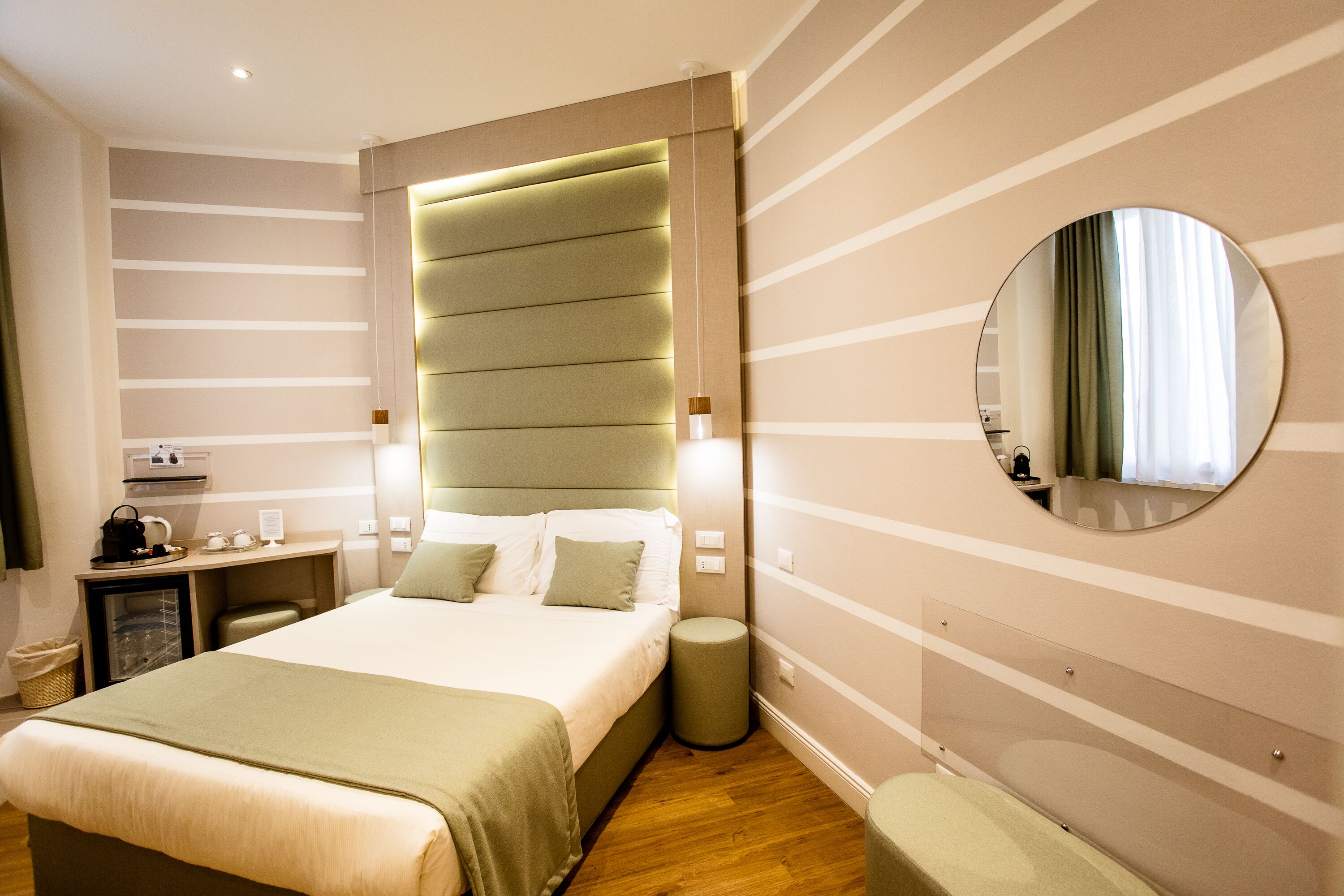 A hotel room with a large bed, bedside tables, a round mirror, and a small stool.